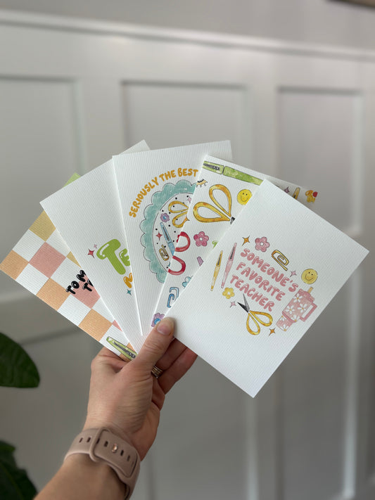 Hand holding a collection of greeting cards with various designs against a white paneled wall.