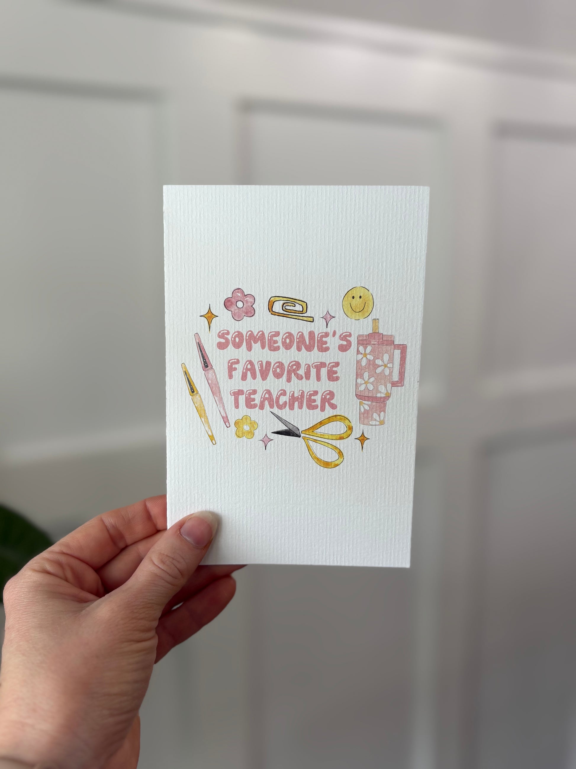 Hand holding a card with 'Someone's Favorite Teacher' text and illustrations on a neutral background