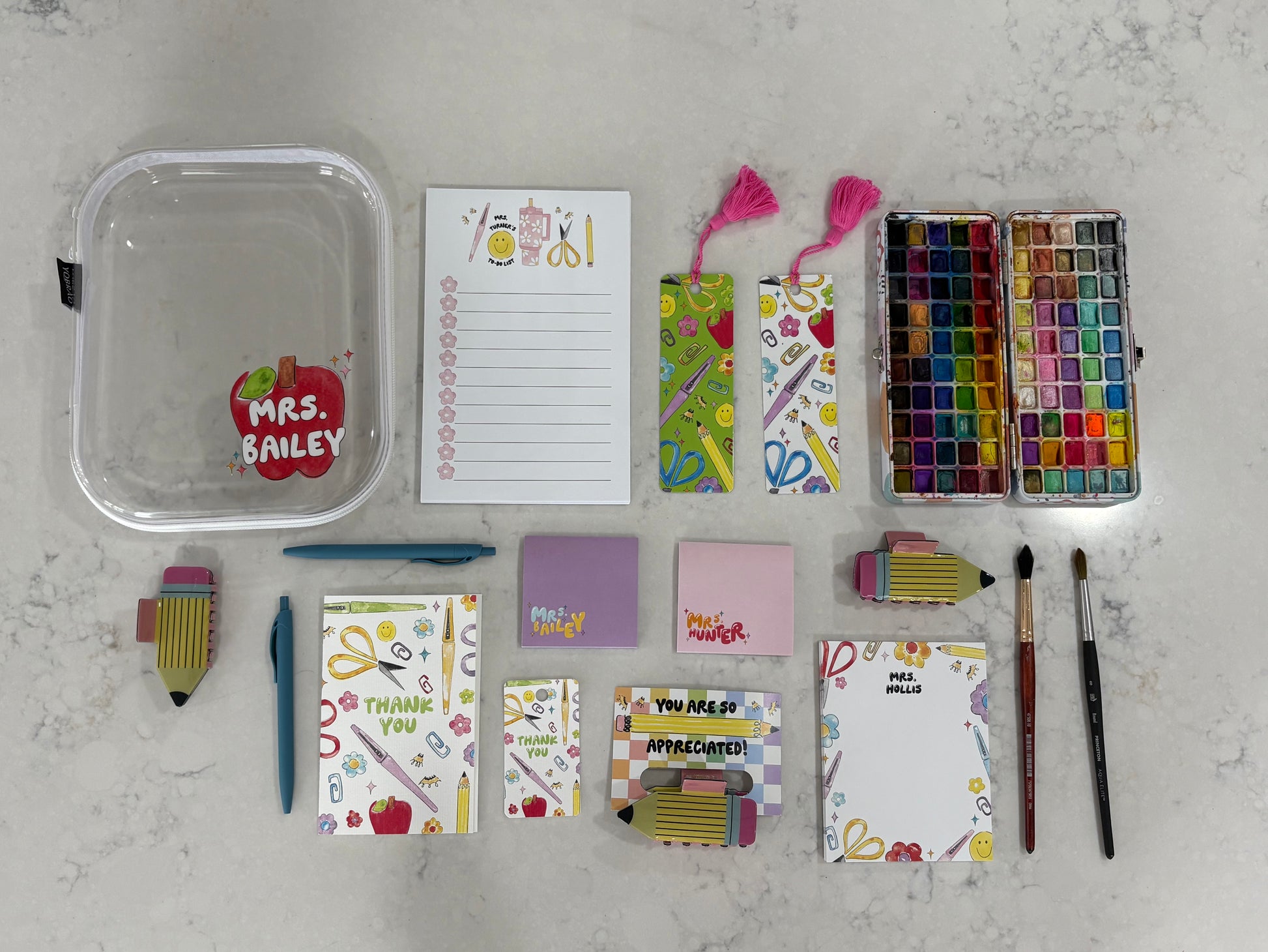 Collection of stationery items including notepads, paint supplies, and a clear container on a light gray surface.