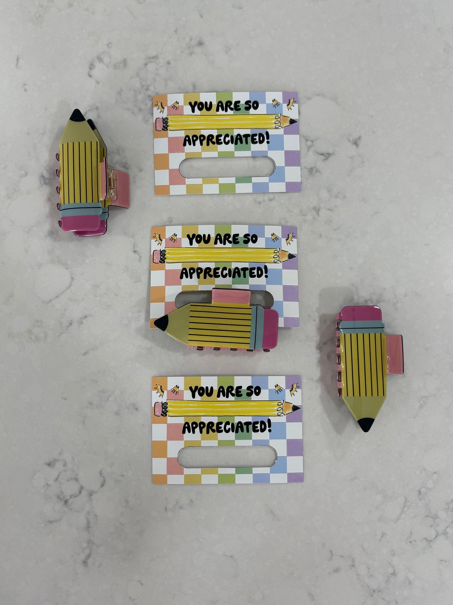 Pencil hair clips held by a colorful holder with 'You are so appreciated!' text on a marble surface