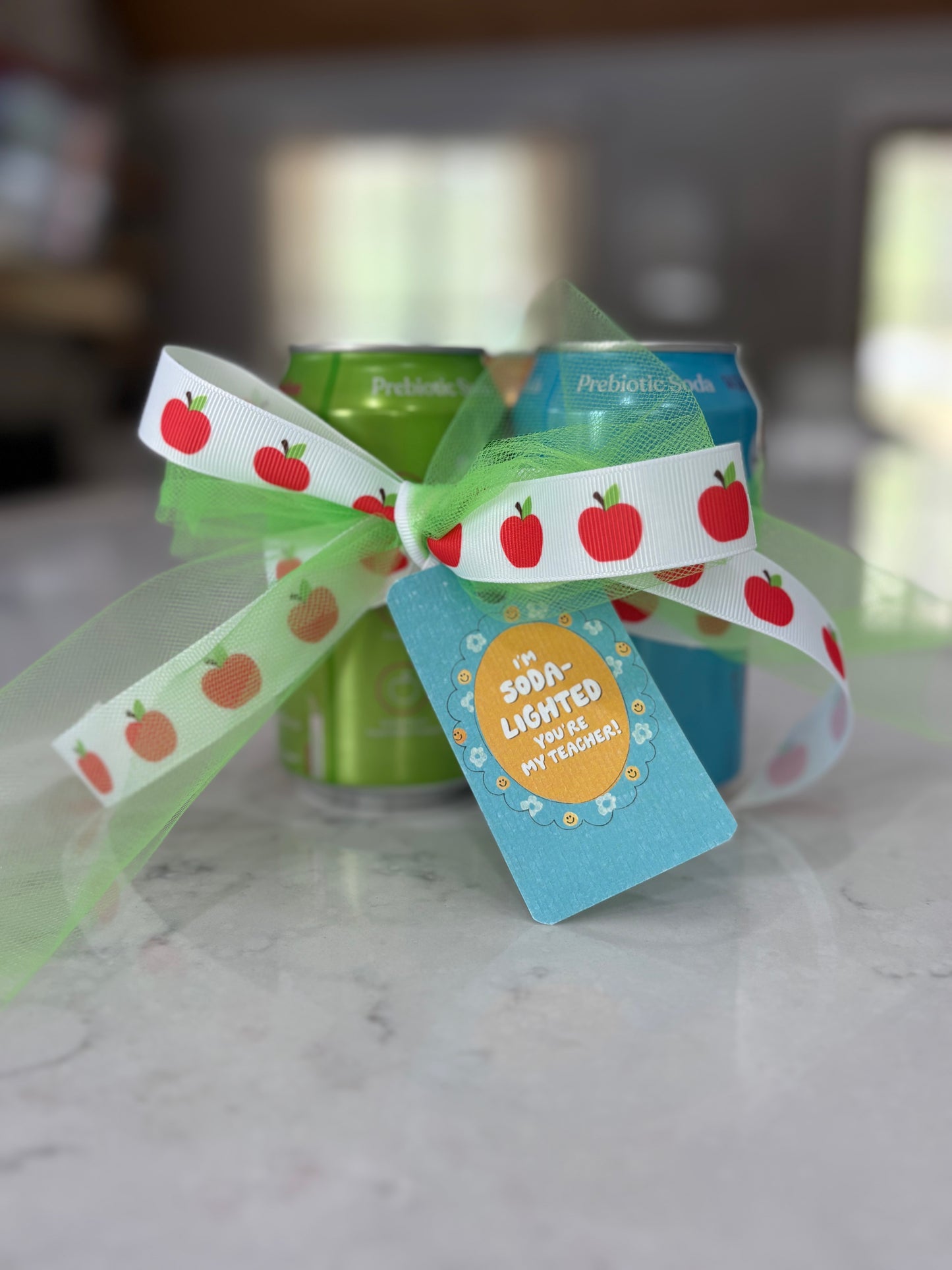 Two cans of soda with a decorative ribbon and tag on a marble surface.