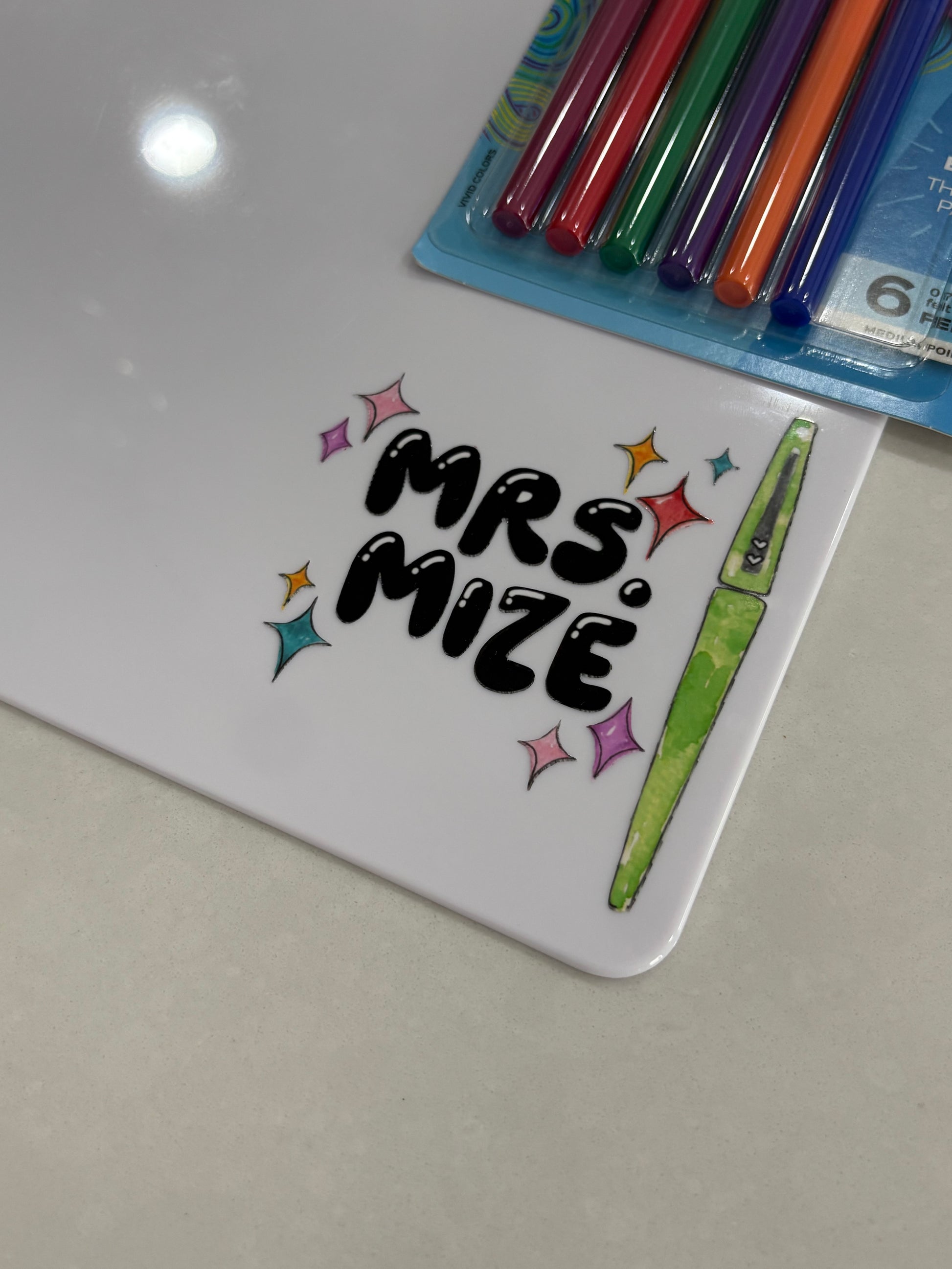 Clipboard with 'Mrs. Mize' on it next to colored pens on a light gray background