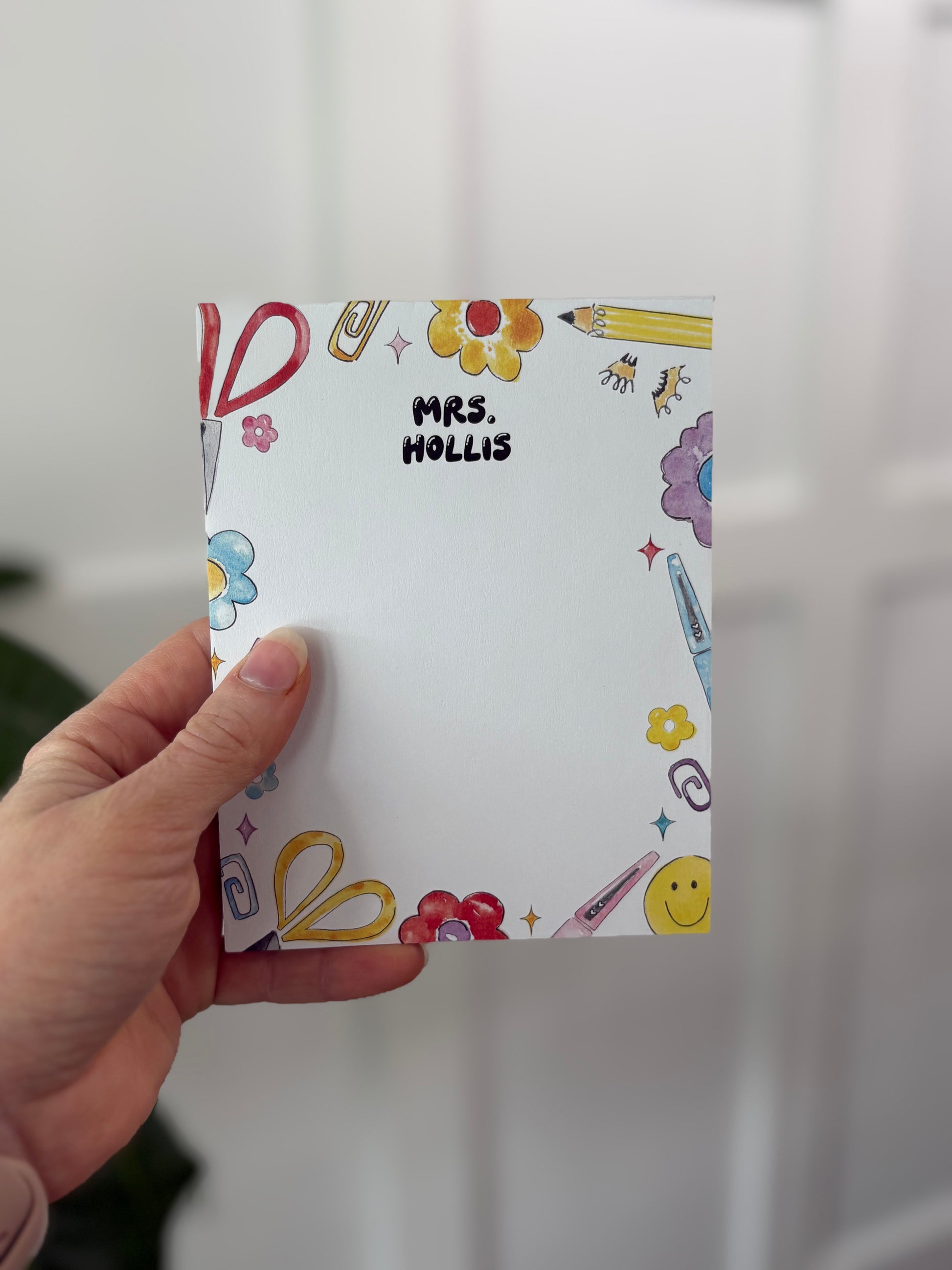 Hand holding a colorful notepad with 'MRS. HOLLIS' printed on it against a white background