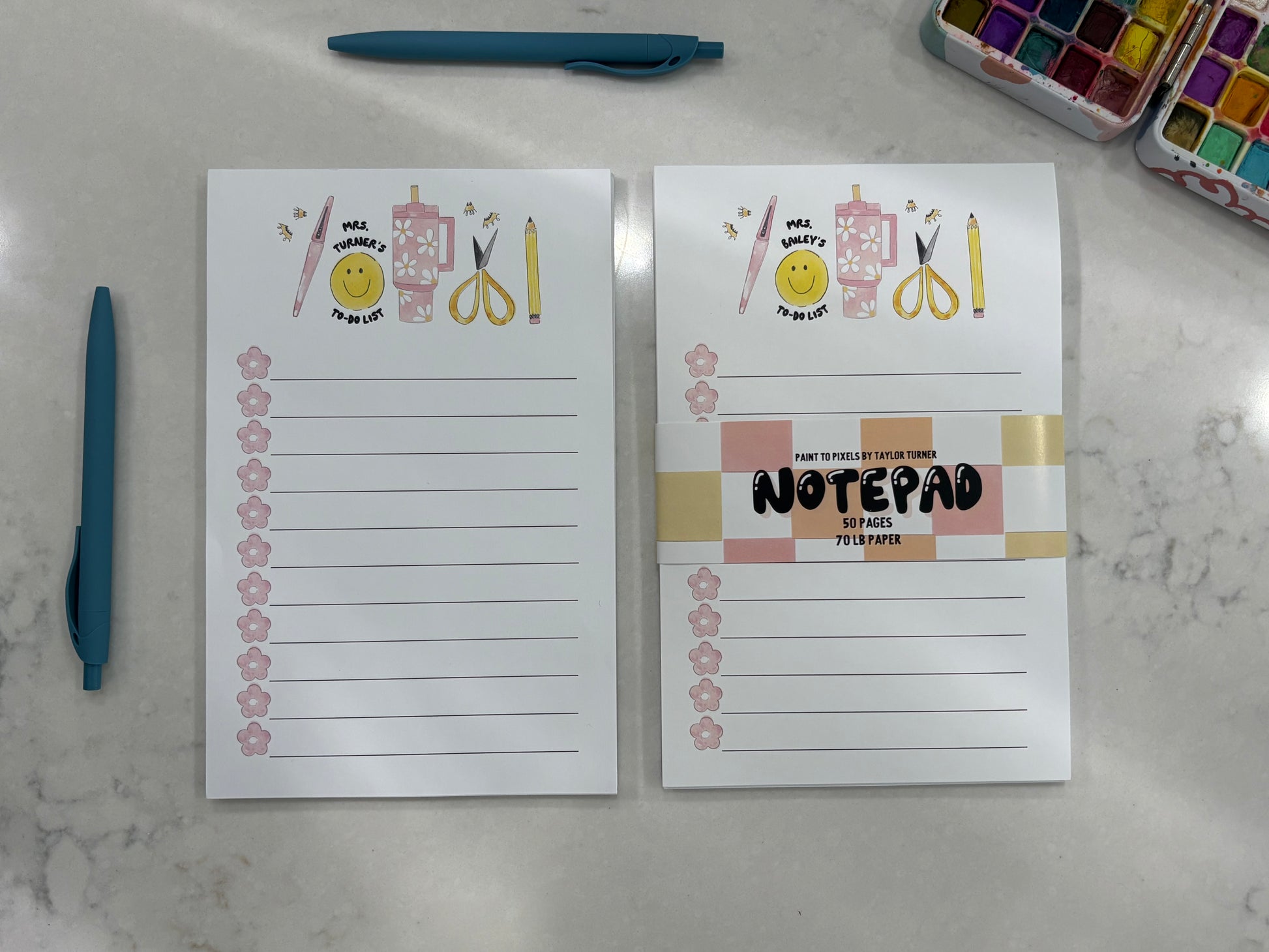 Two notepads with colorful designs on a marble surface with pens and watercolor paint