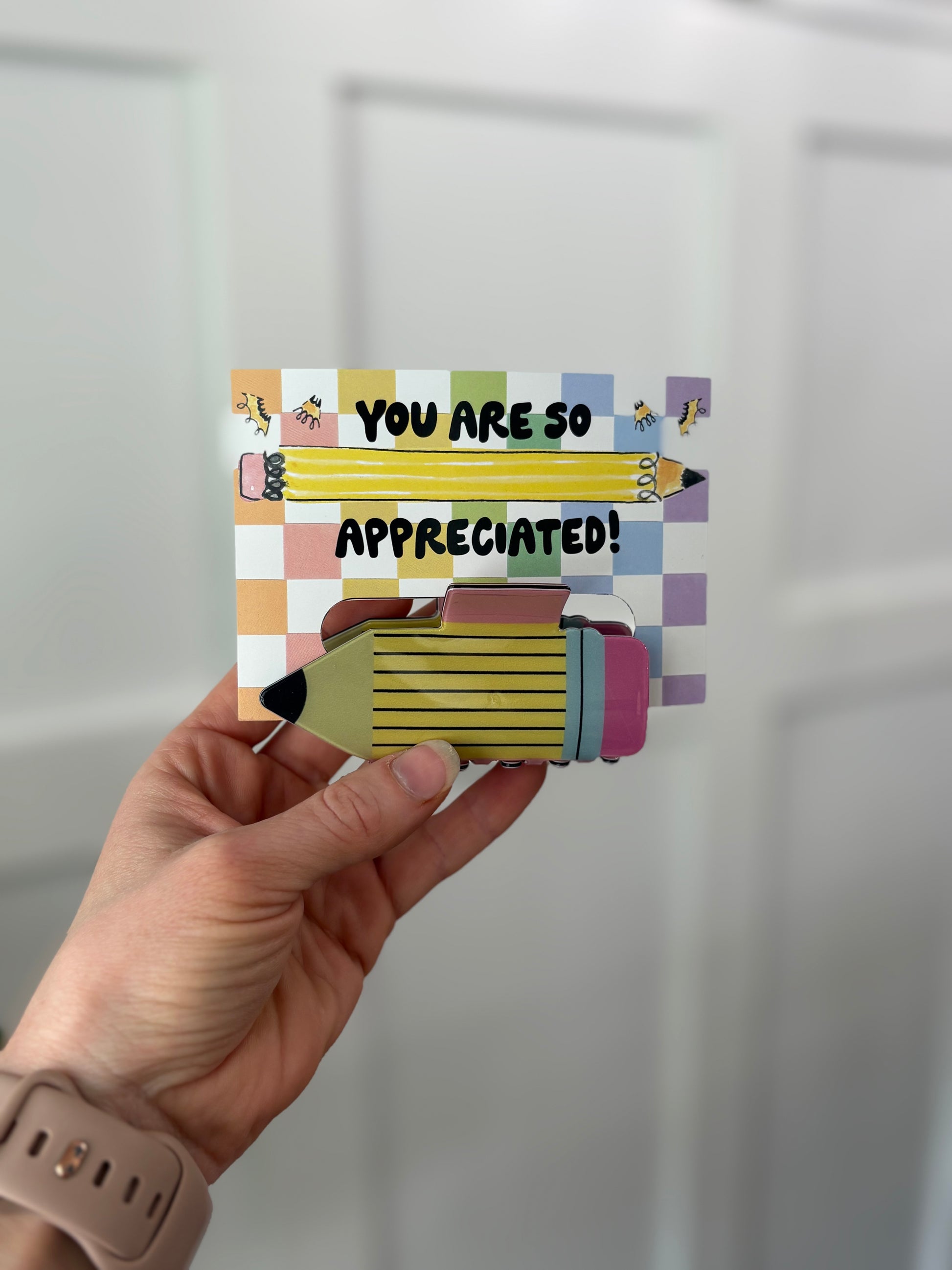 Hand holding a colorful card with 'You are so appreciated!'  text holding a pencil hair clip against a light gray background