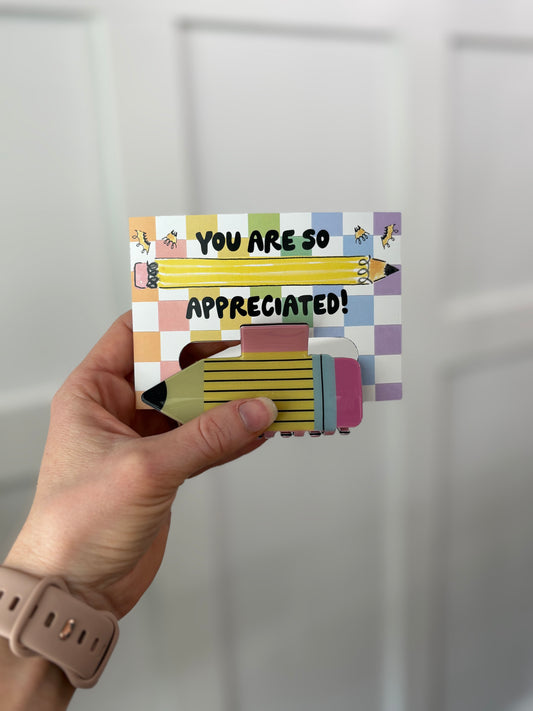 Hand holding a colorful card with 'You are so appreciated!' text holding a hair clip against a white background