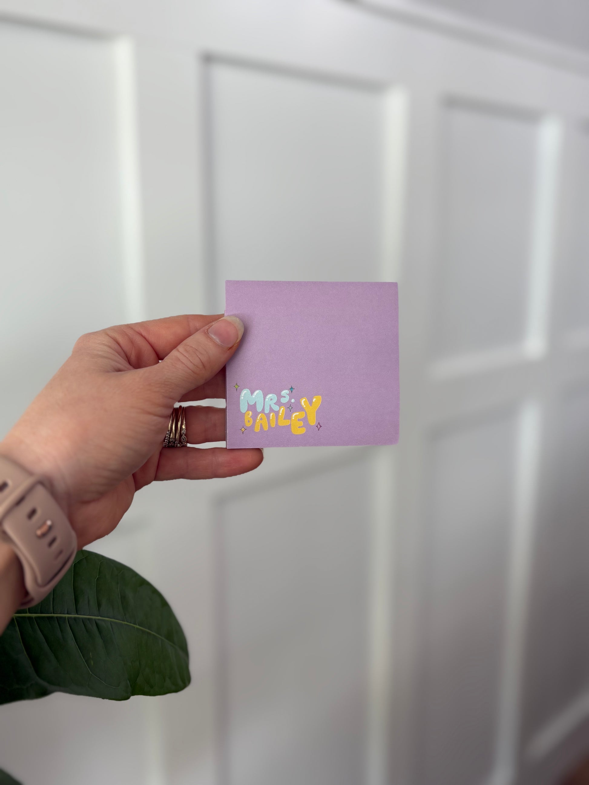 Hand holding purple sticky note pad with 'MRS. Bailey' printed on it against a white background
