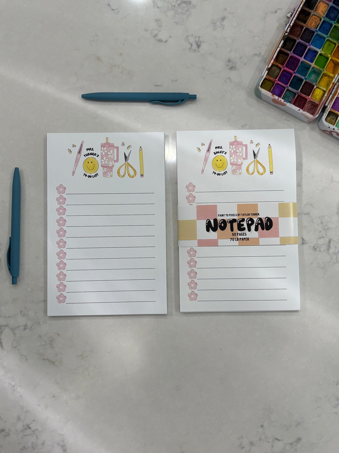 Two notepads with decorative designs on a marble surface with pens and watercolor sets.