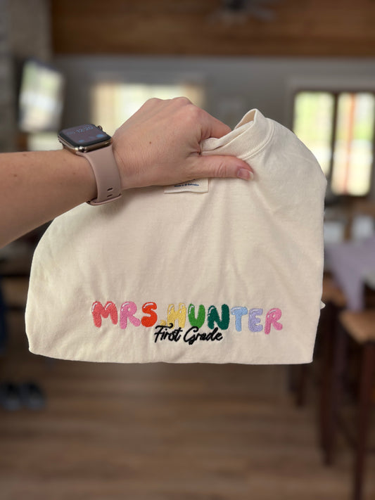 White shirt with 'Mrs. Hunter First Grade' text held by a person indoors.