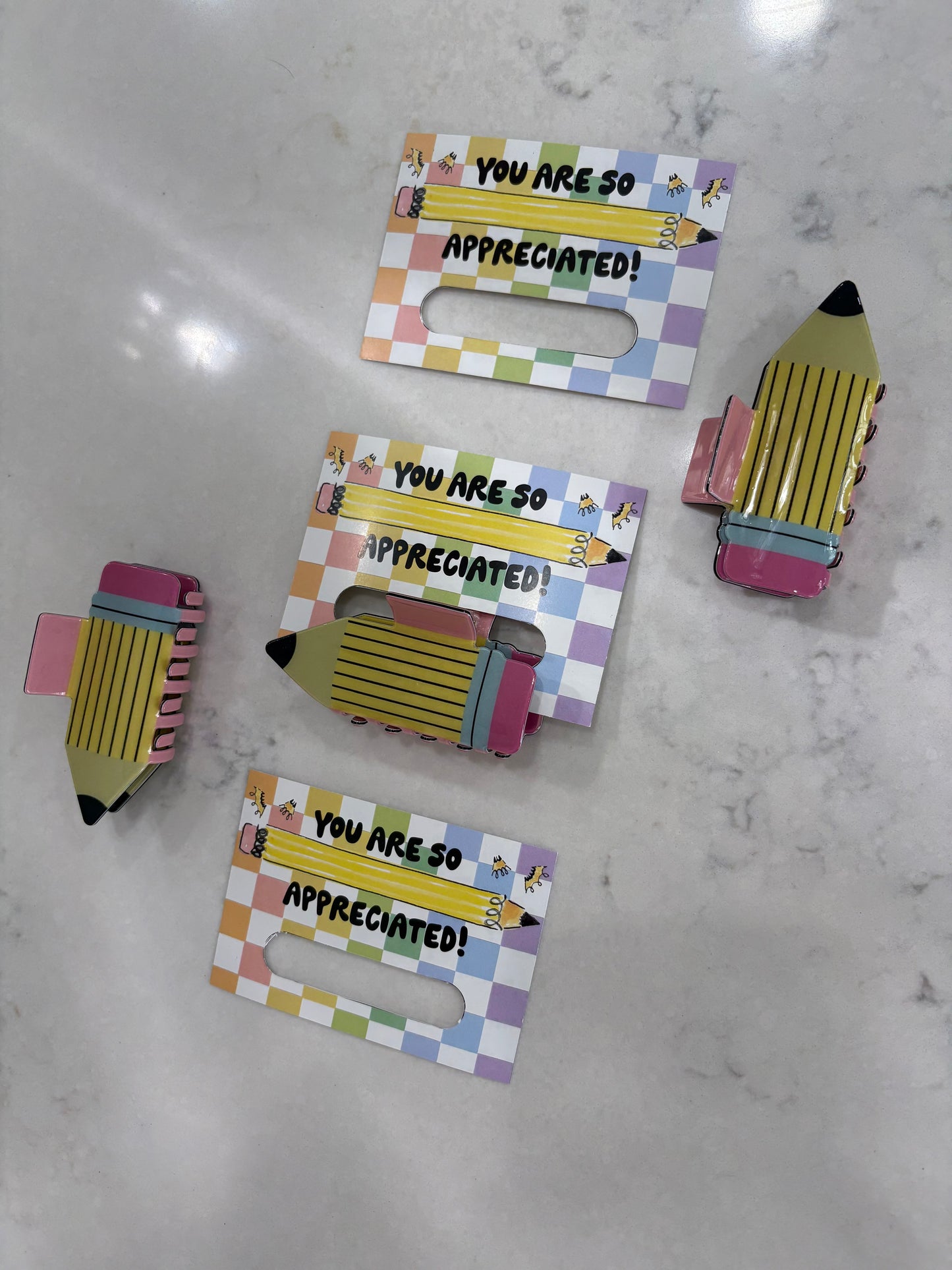 Pencil hair clips held by a colorful holder with 'You are so appreciated!' text on a marble surface