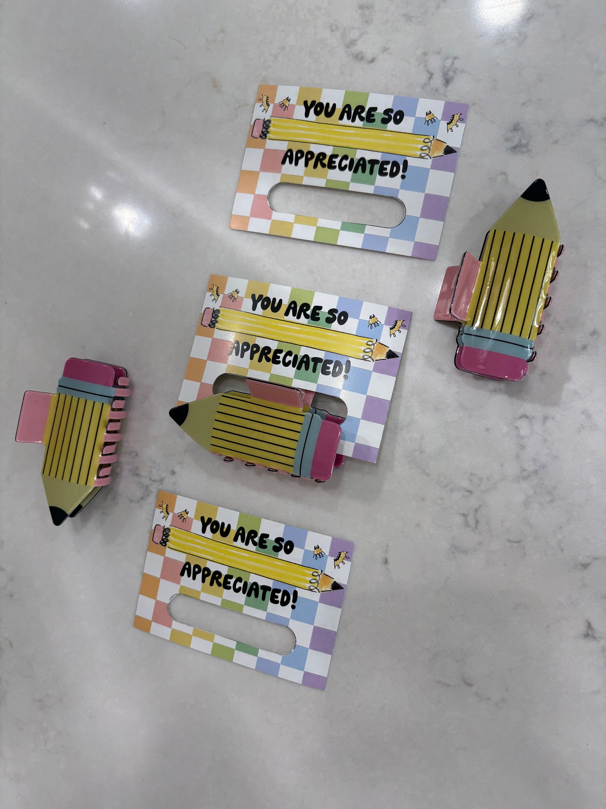 Pencil hair clips held by a colorful holder with 'You are so appreciated!' text on a marble surface
