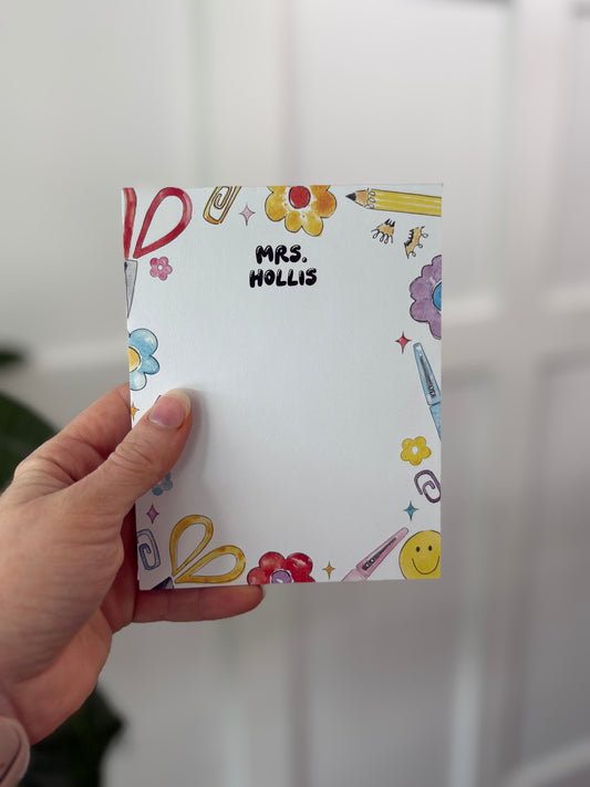 Hand holding a colorful notepad with 'MRS. HOLLIS' printed on it against a white background