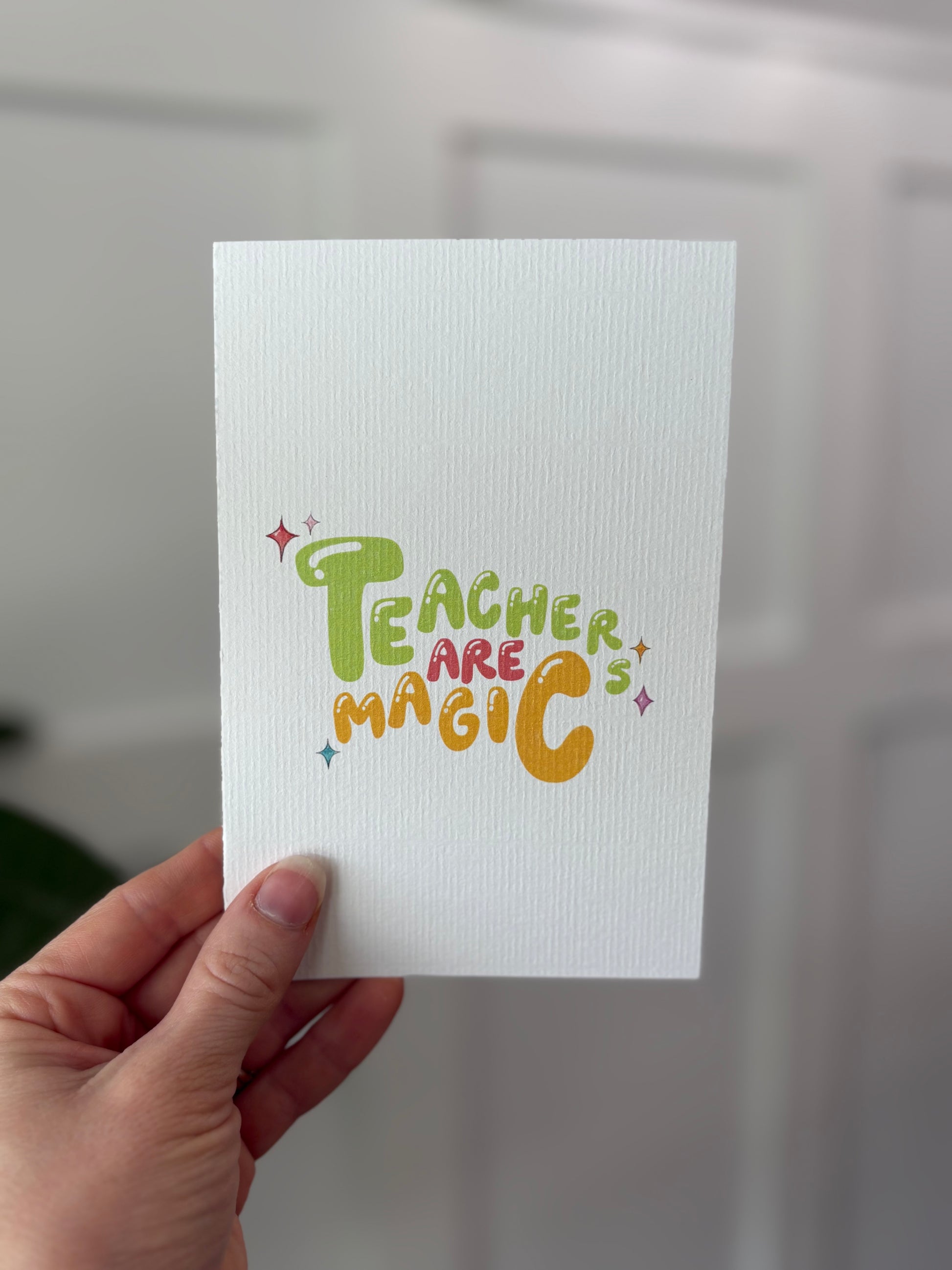Hand holding a card with 'Teachers are Magic' text in colorful letters against a neutral background.
