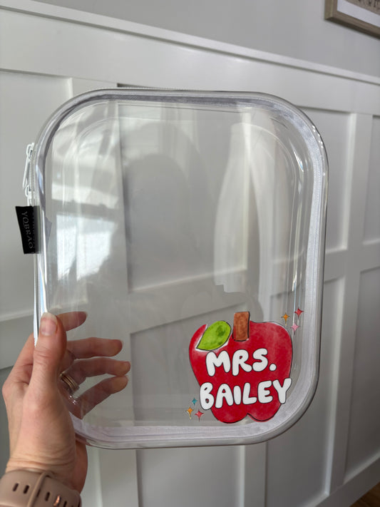 Clear plastic container with a 'Mrs. Bailey' inside of a red apple in the bottom-right corner. Held by a hand against a white surface.