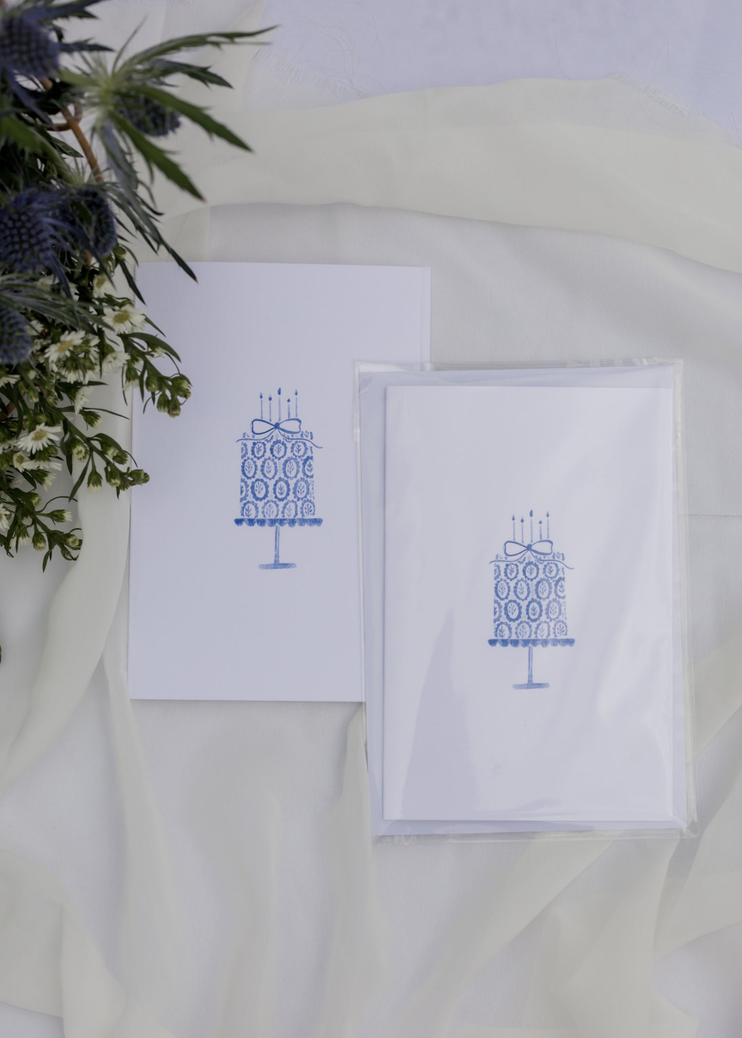 Two white cards with blue cake illustrations on a white surface with greenery.