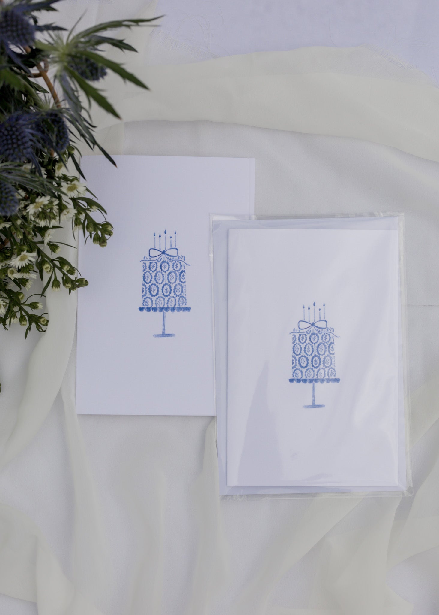 Two white cards with blue cake illustrations on a white surface with greenery.