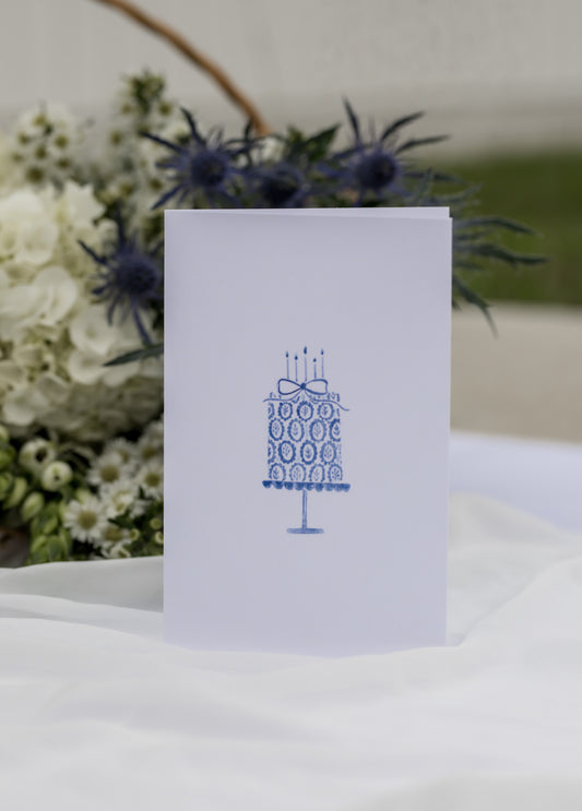 White card with blue cake design on a white surface with flowers in the background