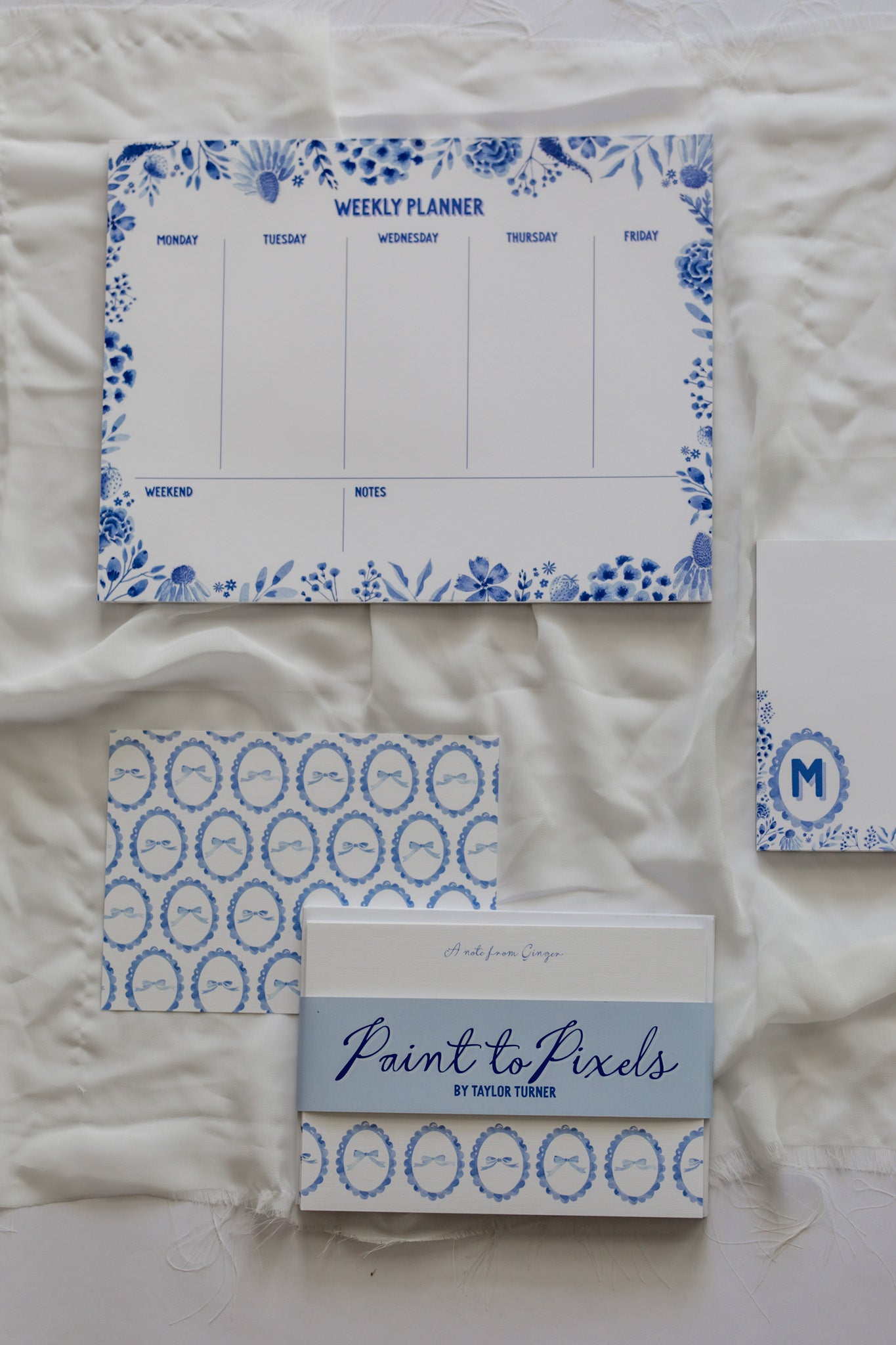 Bow patterned stationery cards wrapped with a branding sleeve labeled "Paint to Pixels," A floral weekly calendar, and a "M" initial memo pad with a white fabric background. 
