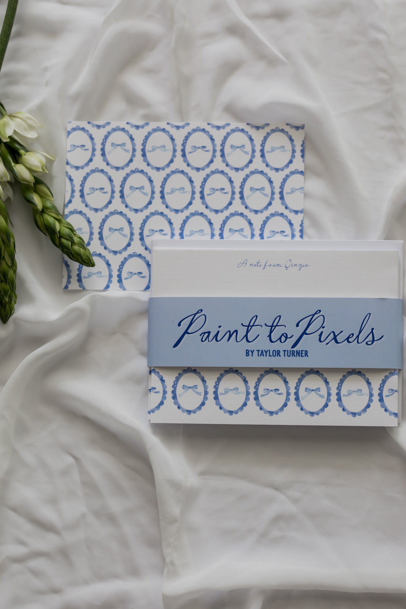 Bow patterned stationery cards wrapped with a branding sleeve labeled "Paint to Pixels" with a white fabric background and a flower arrangement. 