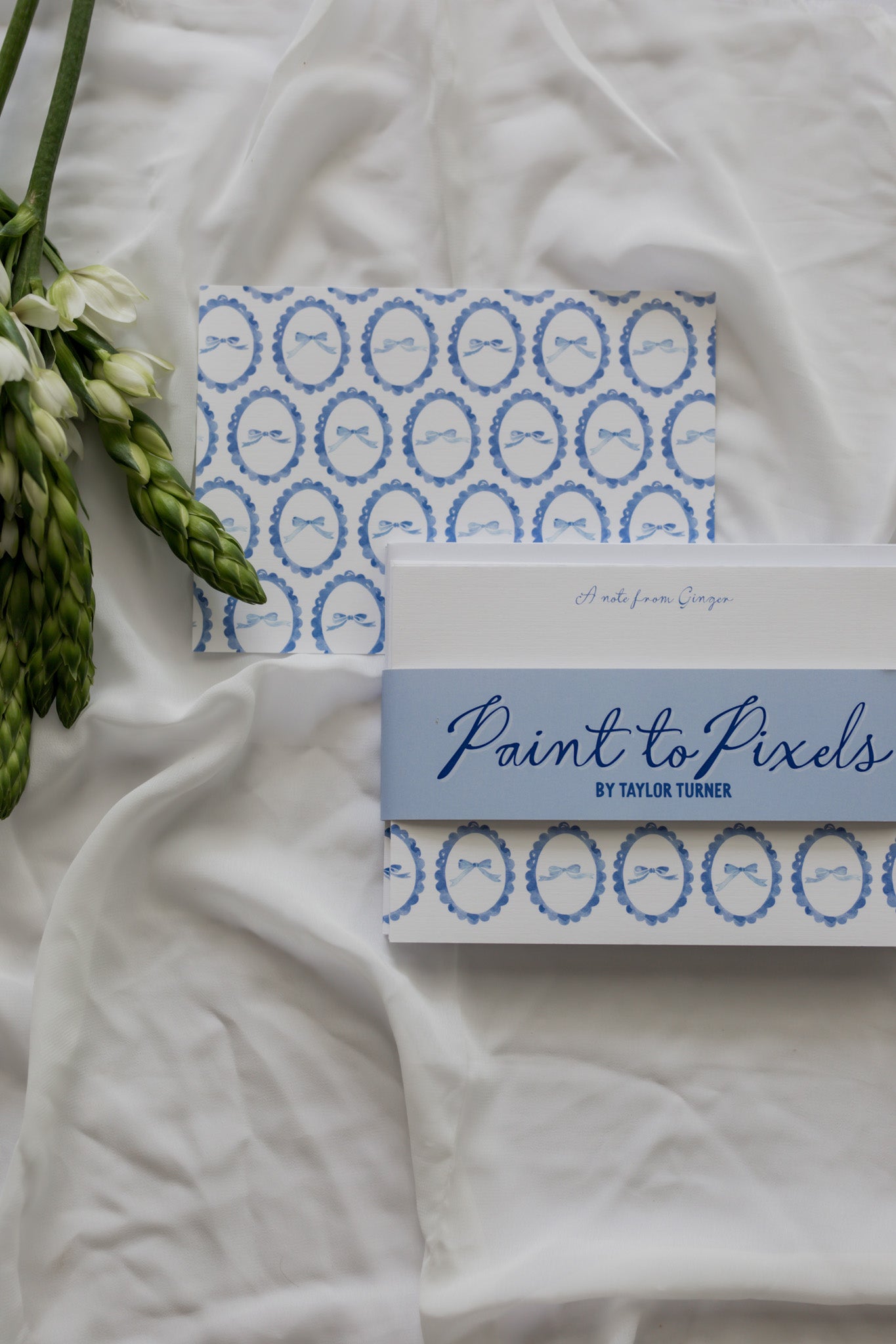 Bow patterned stationery cards wrapped with a branding sleeve labeled "Paint to Pixels" with a white fabric background and a flower arrangement. 