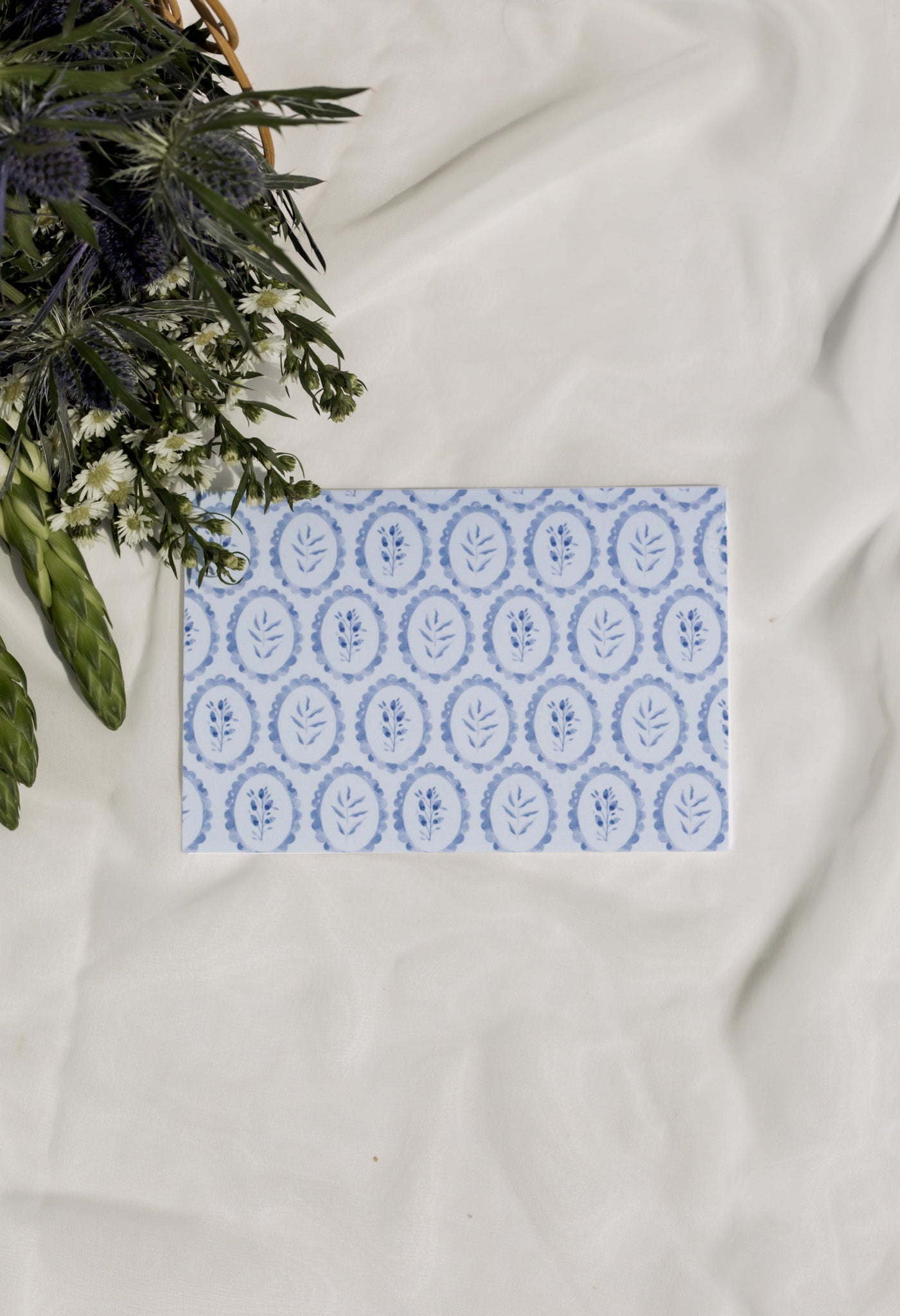 Card with blue floral pattern on a white surface with flowers in the background