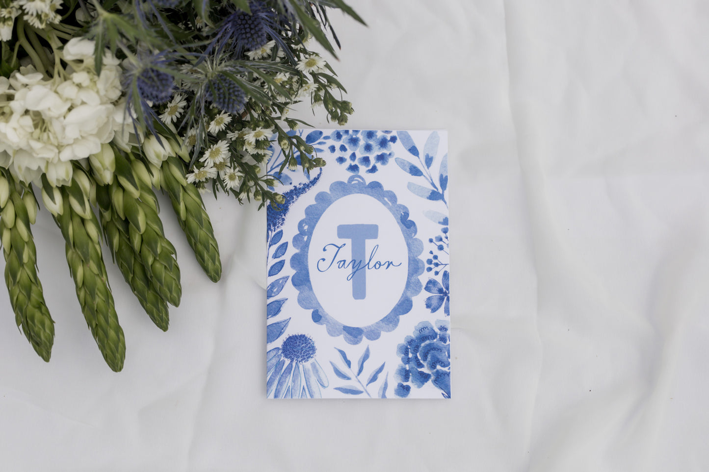 Personalized card with blue floral design and 'Taylor' monogram on a white surface with greenery.