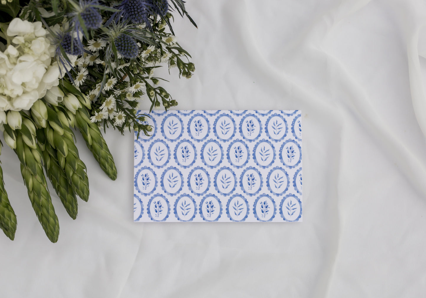 Card with blue floral pattern on a white surface with flowers in the background