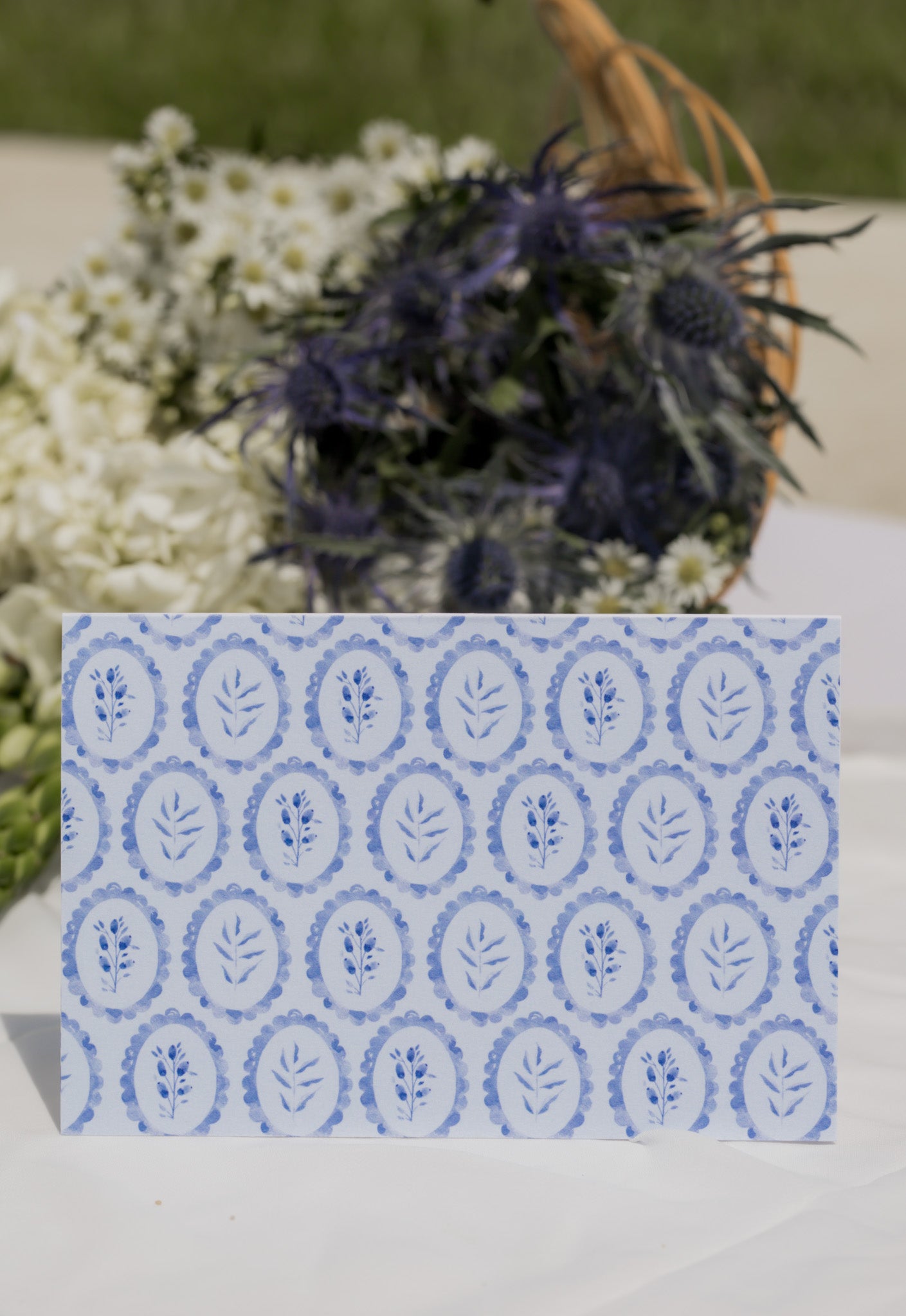 Card with blue floral pattern on a white surface with flowers in the background