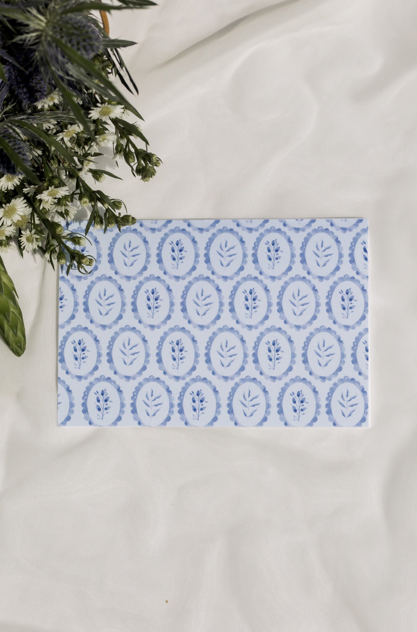 Card with blue floral pattern on a white surface with flowers in the background