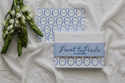 Bow patterned stationery cards wrapped with a branding sleeve labeled "Paint to Pixels" with a white fabric background and a flower arrangement. 