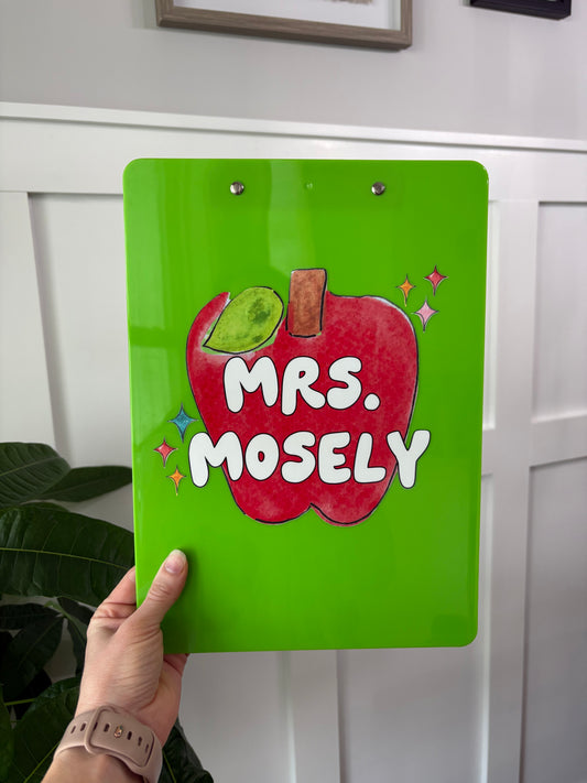 Green clipboard with red apple and 'Mrs. Mosely' text held by a hand against a white door.