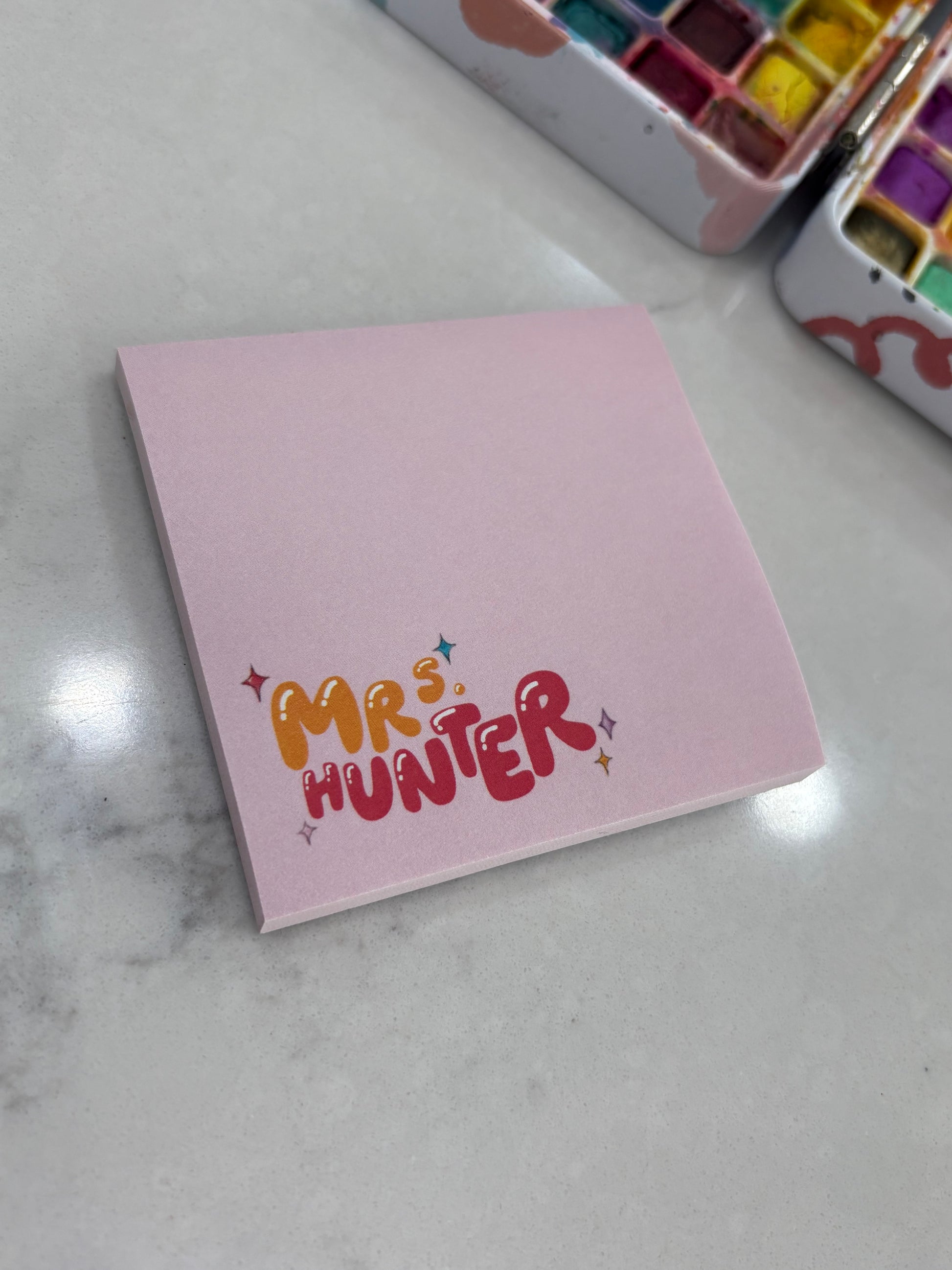 Pink notebook with 'Mr. & Mrs. Hunter' on a marble surface with watercolor sets.