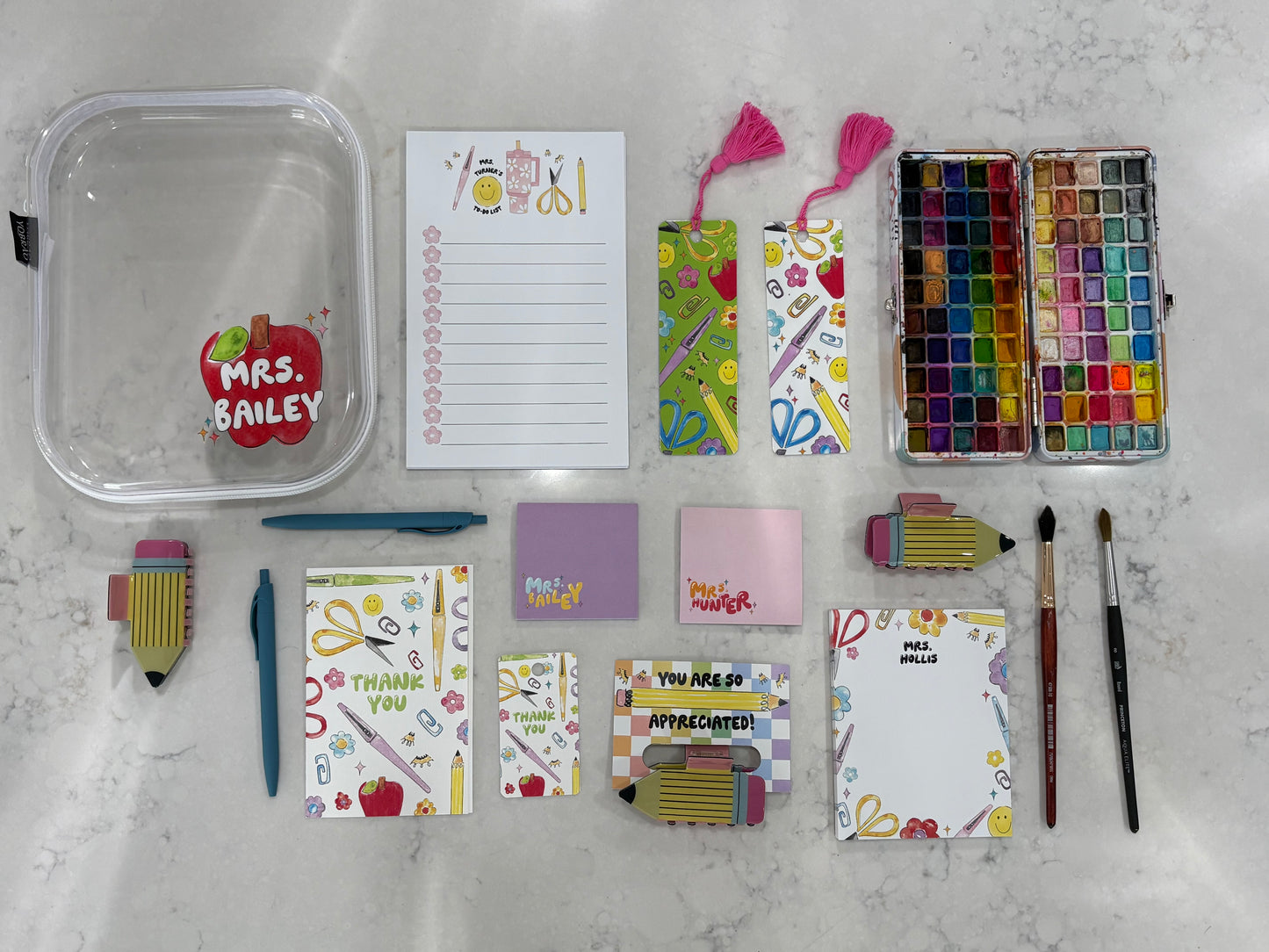 Collection of stationery items including notepads, bookmarks, sticky notes, hair clips, pens, paint brushes, paint, and a container on a marble surface.