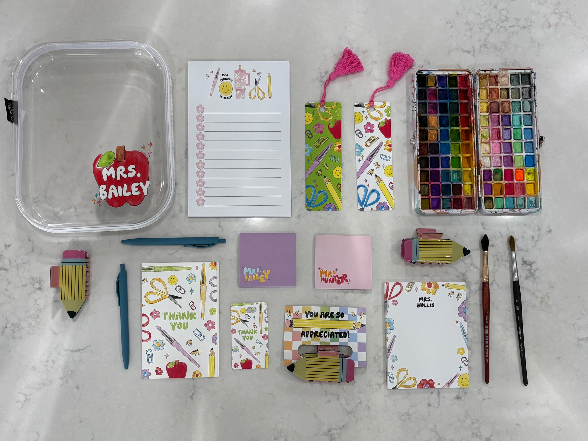 Collection of stationery items including notepads, bookmarks, sticky notes, hair clips, pens, paint brushes, paint, and a container on a marble surface.