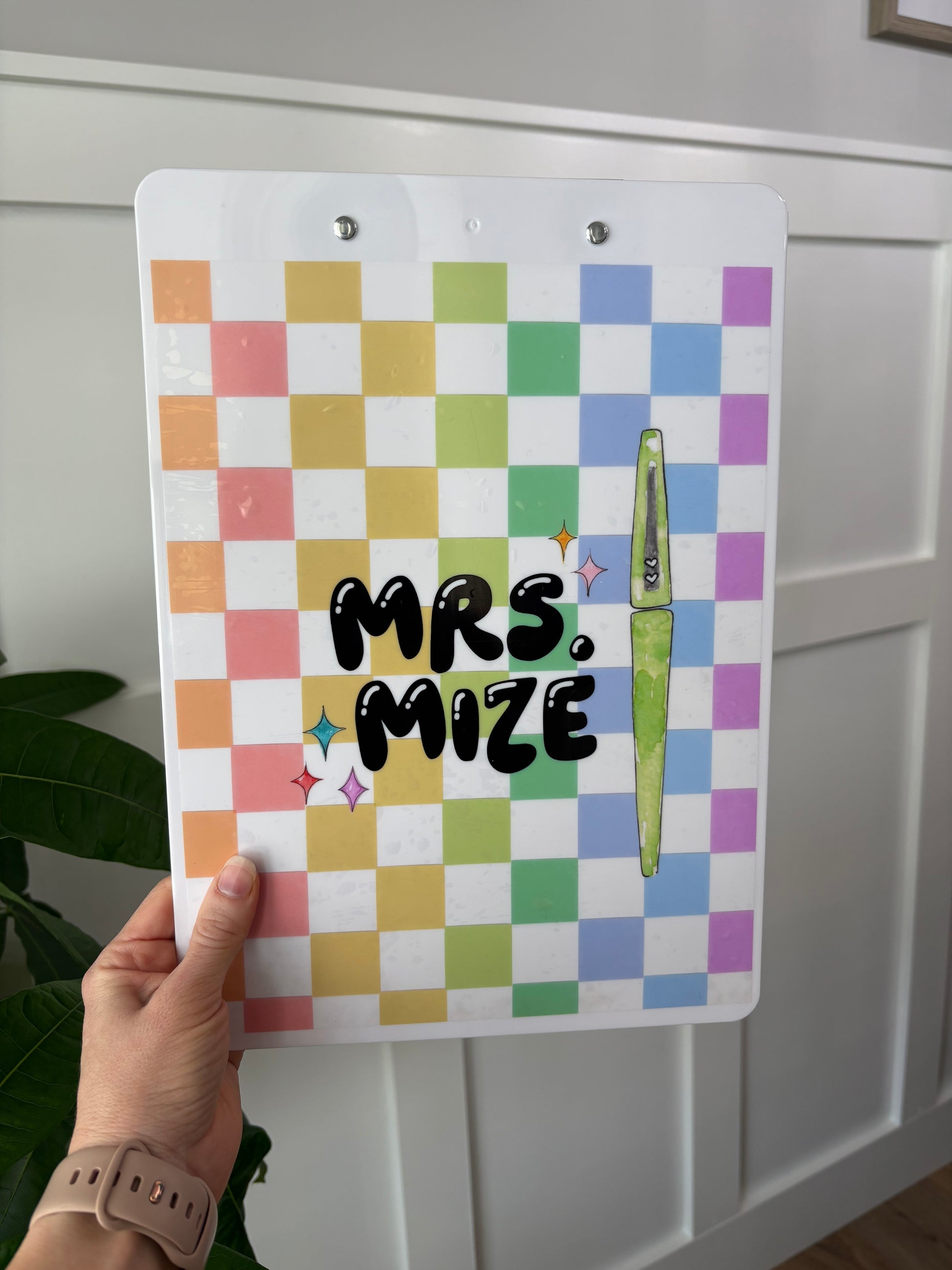 Colorful checkered clipboard with 'Mrs. Maze' text held by a hand against a white background