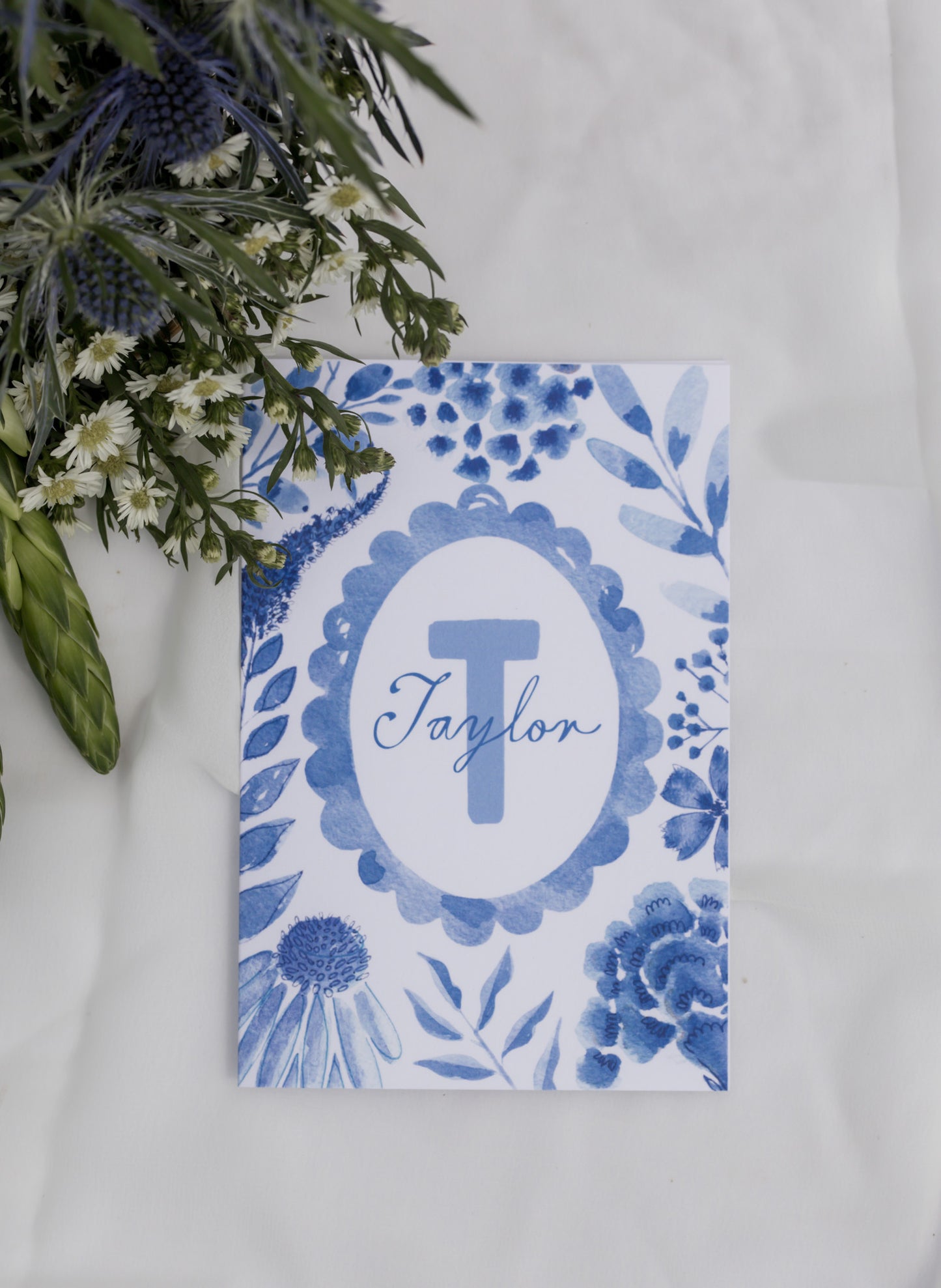 Personalized card with blue floral design and 'Taylor' monogram on a white surface with greenery.