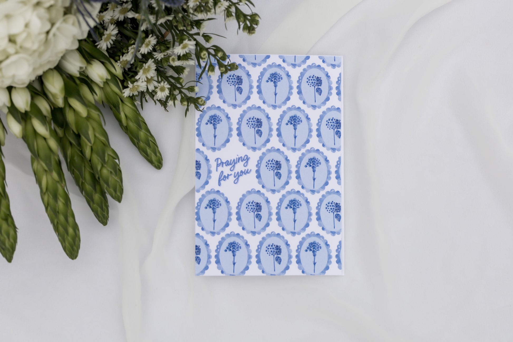 Greeting card with blue floral pattern and text next to asparagus and flowers on a white surface