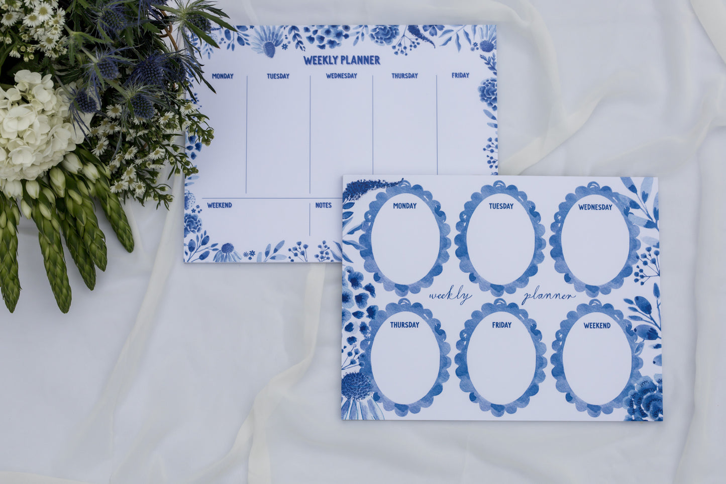 Two Weekly planners with blue floral designs on a white surface with flowers.