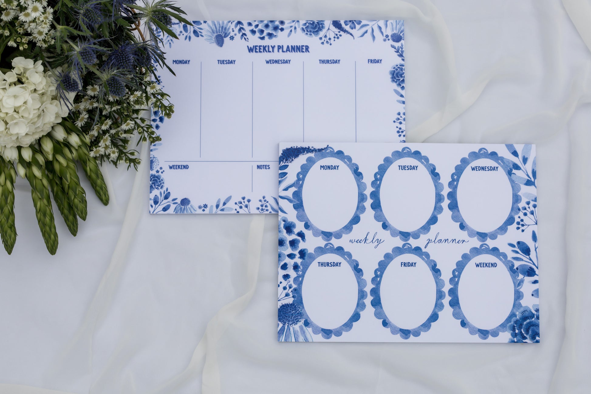 Two Weekly planners with blue floral designs on a white surface with flowers.