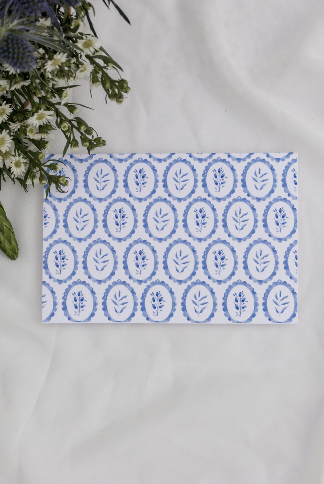 Card with blue floral pattern on a white surface with flowers in the background