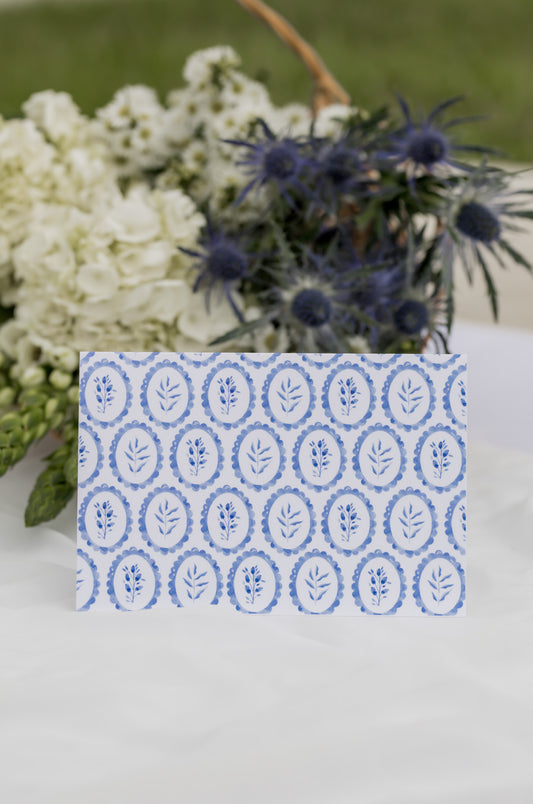 Card with blue floral pattern on a white surface with flowers in the background