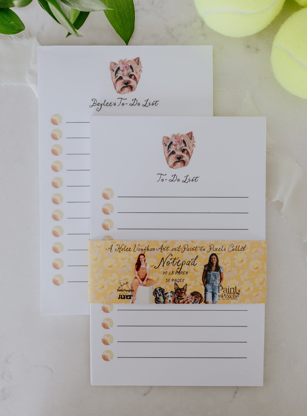 A stack of two To-Do List with lines, tennis balls, and a dog portrait featuring a Shih Tzu. Wrapped in a yellow belly band with styled with pictures of the artists, dogs, and branding. All sitting on a marble surface with greenery and tennis balls