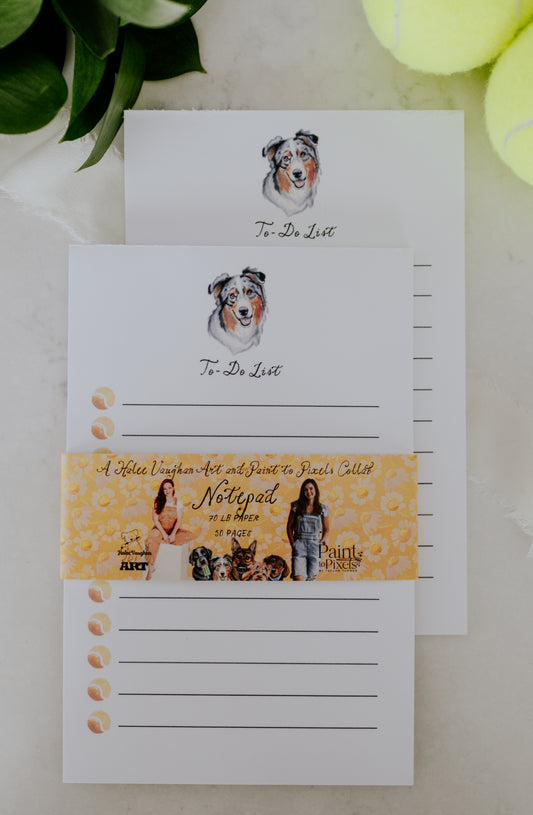 A stack of two To-Do List with lines, tennis balls, and a dog portrait featuring an Australian Shepherd. Wrapped in a yellow belly band with styled with pictures of the artists, dogs, and branding. All sitting on a marble surface with greenery and tennis balls