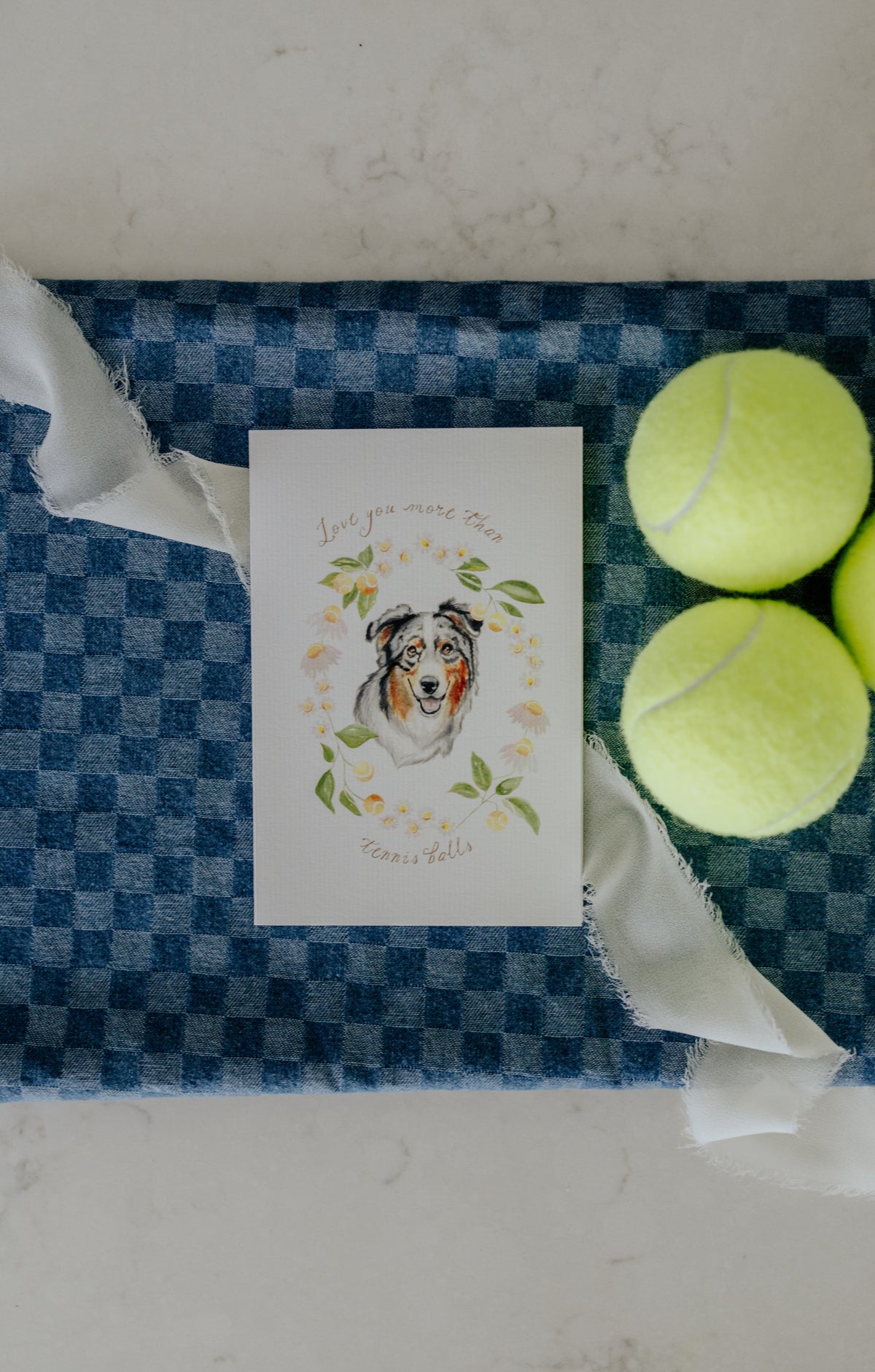 A square white greeting card featuring a watercolor illustration of a dog surrounded by flowers and the words 'Love you more than tennis balls,' styled on a blue-and-white checkered towel with two tennis balls beside it.