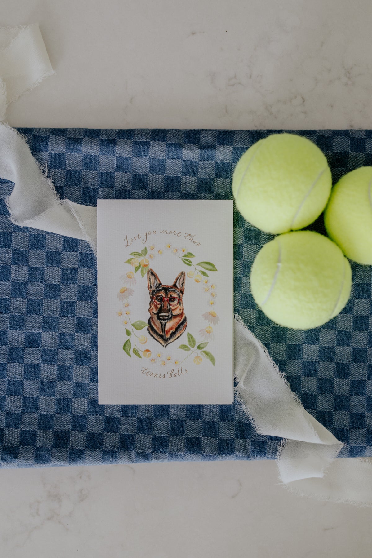 A square greeting card featuring a watercolor illustration of a German Shepherd dog surrounded by green leaves and small yellow flowers, with the text 'Love you more than tennis balls.' The card is placed on a blue-and-white checkered fabric surface, styled with three yellow tennis balls and white ribbon on a light marble-like background.