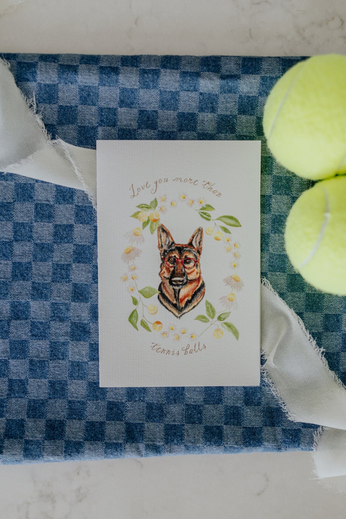A square white greeting card featuring a watercolor illustration of a German Shepherd dog surrounded by leaves and small flowers, with the text 'Love you more than Kennel Falls' above and below. The card is placed on a blue-and-white checkered cloth with white ribbon and two yellow tennis balls nearby.