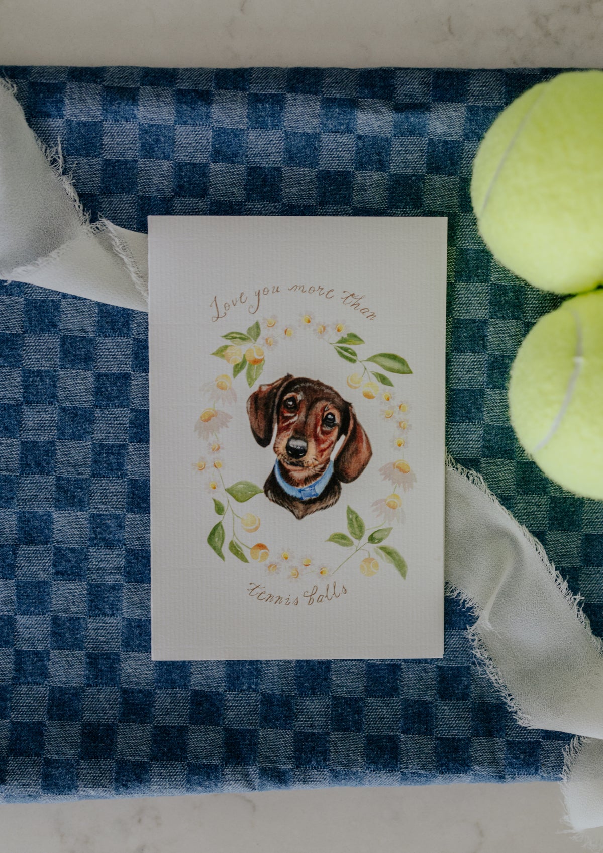 A square white greeting card featuring a watercolor illustration of a dachshund surrounded by green leaves and yellow tennis balls, with the text 'Love you more than Tennis Balls'. The card is styled on a blue and white checkered fabric alongside two yellow tennis balls and white ribbon.