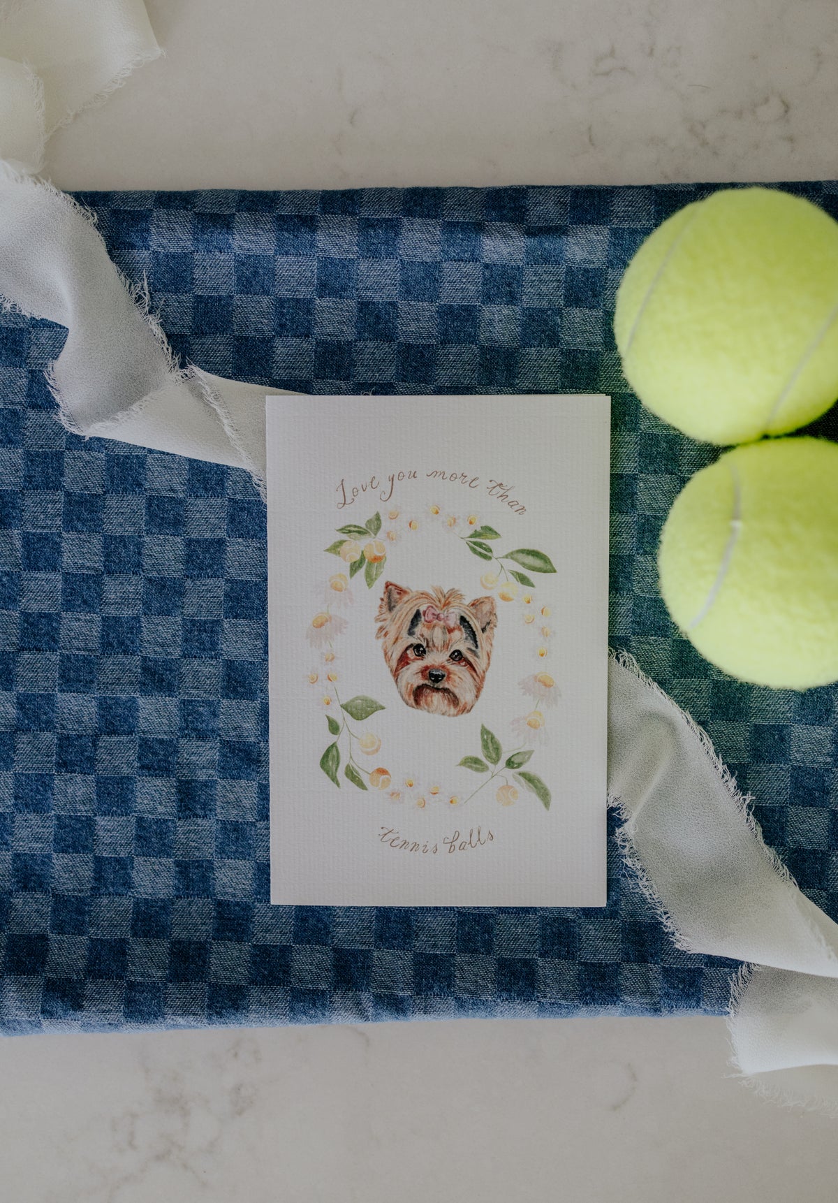 A square white greeting card featuring a watercolor illustration of a Shih Tzu surrounded by green leaves and yellow tennis balls, with the text 'Love you more than Tennis Balls'. The card is styled on a blue and white checkered fabric alongside two yellow tennis balls and white ribbon.