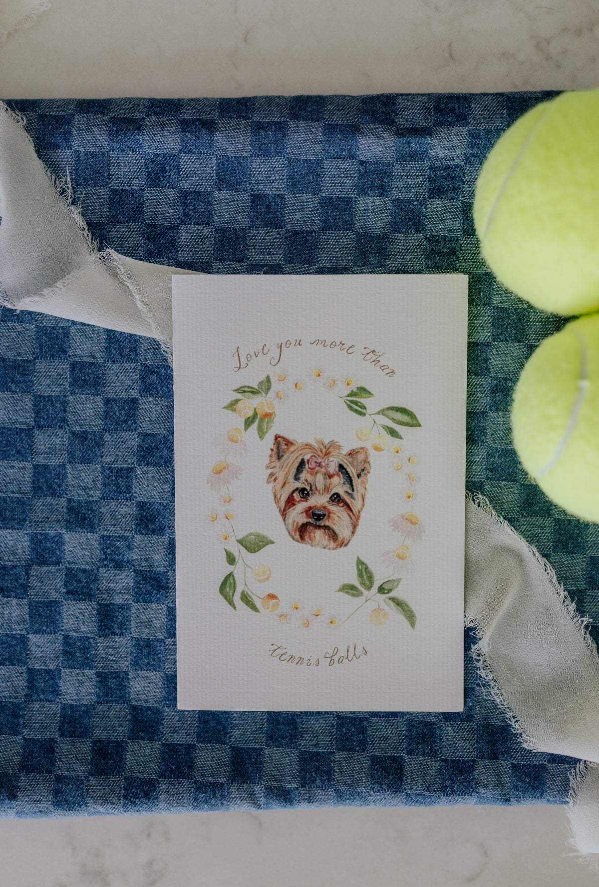 A square white greeting card featuring a watercolor illustration of a shih tzu surrounded by green leaves and yellow tennis balls, with the text 'Love you more than Tennis Balls'. The card is styled on a blue and white checkered fabric alongside two yellow tennis balls and white ribbon.