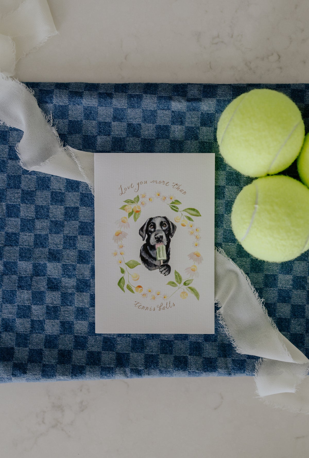 A square white greeting card featuring a watercolor illustration of a black and white dog holding a tennis ball, surrounded by a floral wreath and the text 'Love you more than Tennis Balls'. The card is displayed on a blue and white checkered fabric runner, flanked by two yellow tennis balls on a light marble surface.